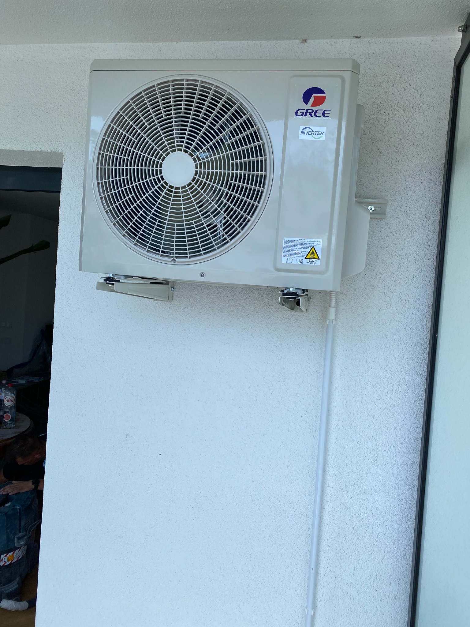 Zewnętrzna jednostka klimatyzatora marki Gree zamontowana na elewacji budynku z widocznymi elementami mocującymi i rurką odprowadzającą skropliny.