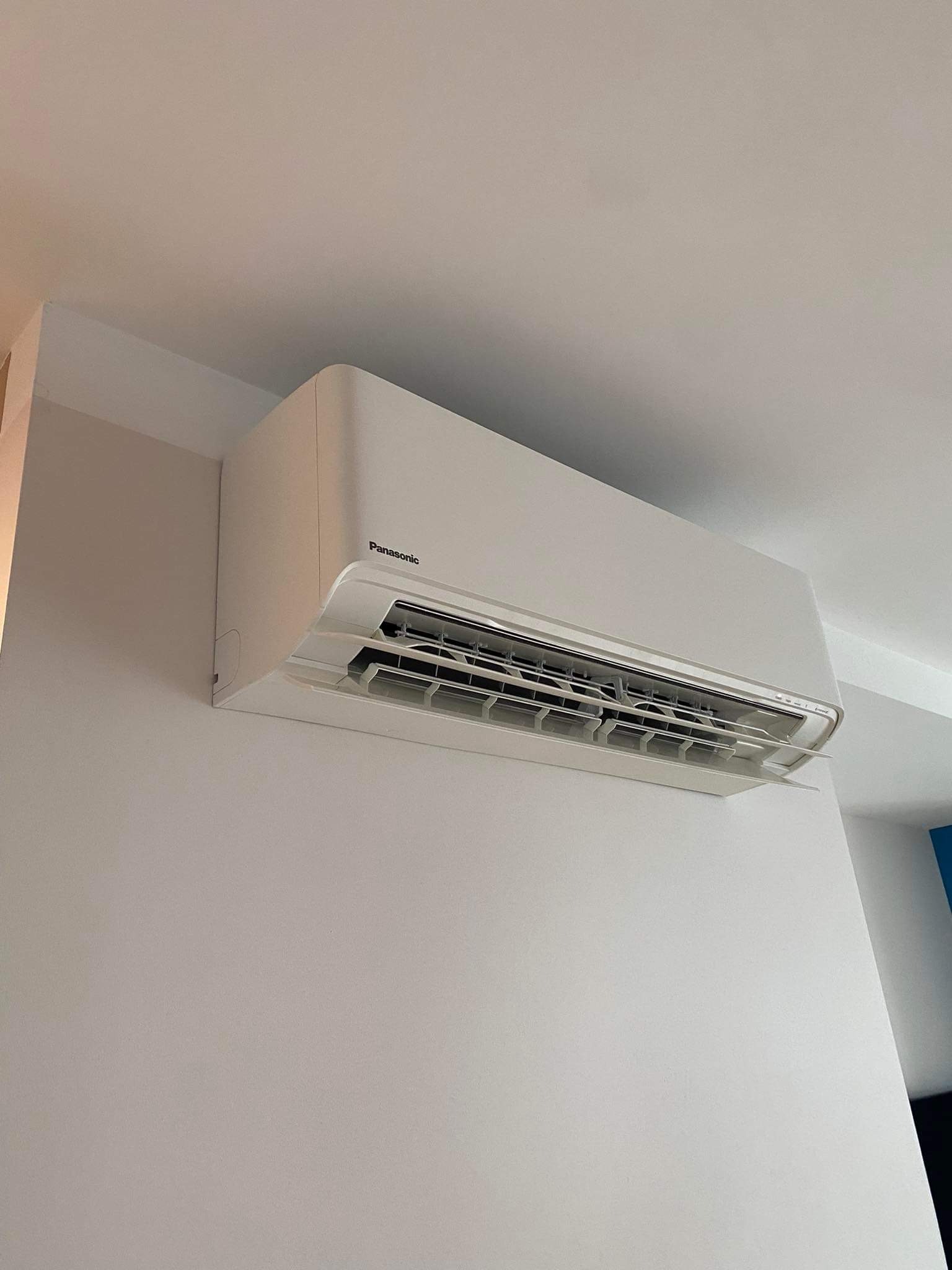 Biała klimatyzacja Panasonic zamontowana na jasnej ścianie, widoczna z dołu, z otwartymi żaluzjami nawiewu.