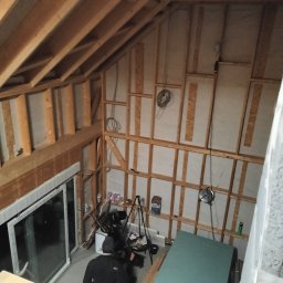 Prywatny wykonawca - Wnętrze poddasza w trakcie adaptacji: drewniana konstrukcja szkieletowa, izolacja między elementami, widoczne przewody elektryczne, okna dachowe i materiały budowlane na podłodze.