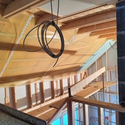 Prywatny wykonawca - Wnętrze poddasza w trakcie adaptacji: widoczne drewniane belki konstrukcyjne, folia paroizolacyjna, fragment komina z szarego betonu komórkowego oraz zwisający kabel elektryczny.