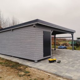 Prywatny wykonawca - Szary, drewniany budynek gospodarczy z czarnymi drzwiami i zadaszeniem, stojący na kostce brukowej, z żółtą skrzynką narzędziową obok, na tle drzew bez liści i pochmurnego nieba.