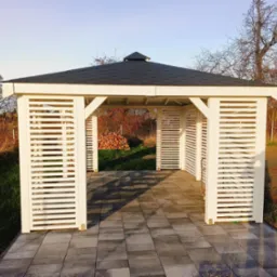 Nowoczesna, biała altana ogrodowa z czarnym dachem, otoczona zielenią i wyłożona szarą kostką brukową, z widocznym stosem drewna wewnątrz.