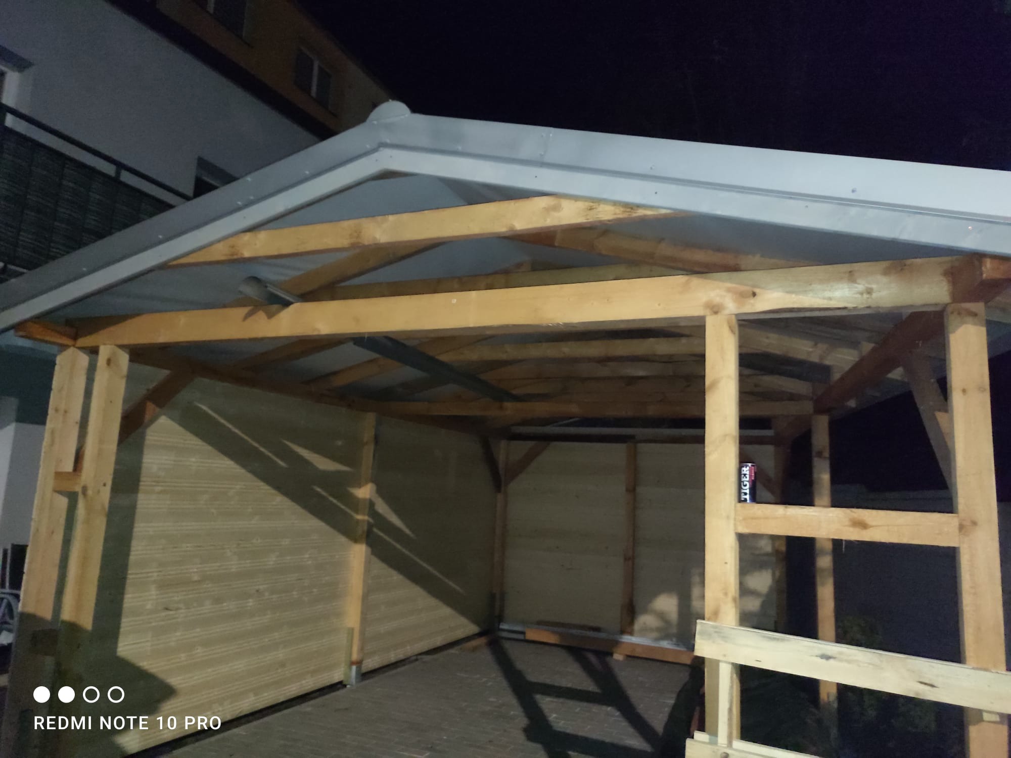 Konstrukcja drewnianej altany w trakcie budowy, widoczne deski ścian i szkielet dachu, puszka napoju energetycznego Tiger na jednej z belek.