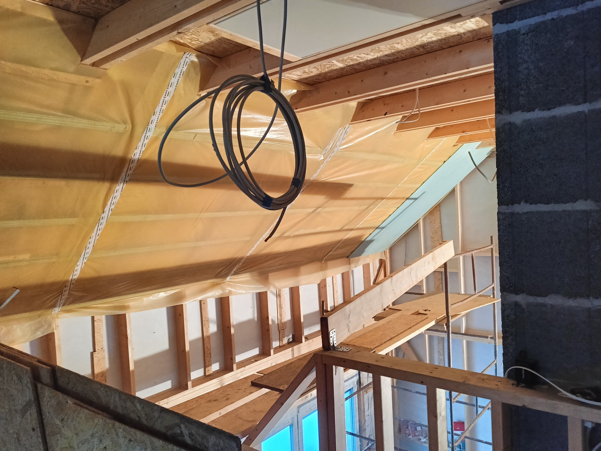 Wnętrze poddasza w trakcie adaptacji: widoczne drewniane belki konstrukcyjne, folia paroizolacyjna, fragment komina z szarego betonu komórkowego oraz zwisający kabel elektryczny.