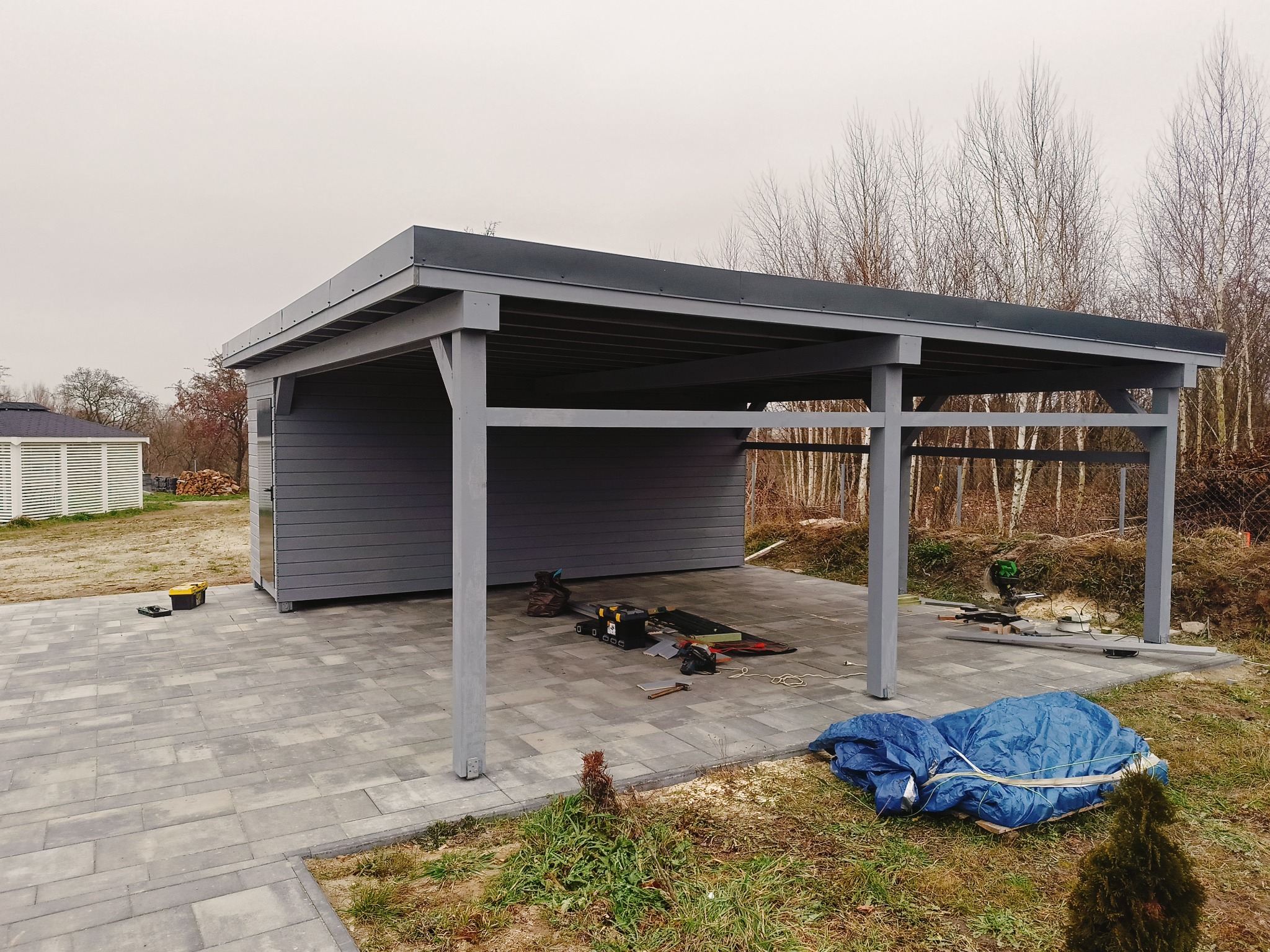 Szara, drewniana wiata garażowa z płaskim dachem, częściowo zabudowana, na posesji z szarą kostką brukową, z narzędziami budowlanymi wewnątrz i niebieską plandeką obok, w tle drzewa bez liści.