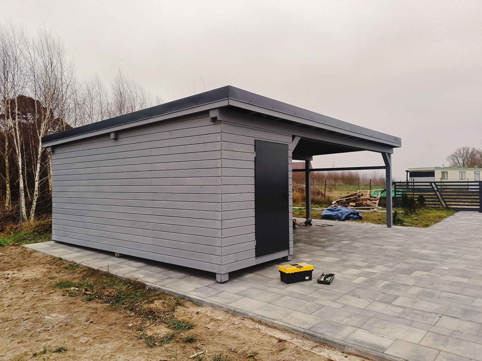 Szary, drewniany budynek gospodarczy z czarnymi drzwiami i zadaszeniem, stojący na kostce brukowej, z żółtą skrzynką narzędziową obok, na tle drzew bez liści i pochmurnego nieba.