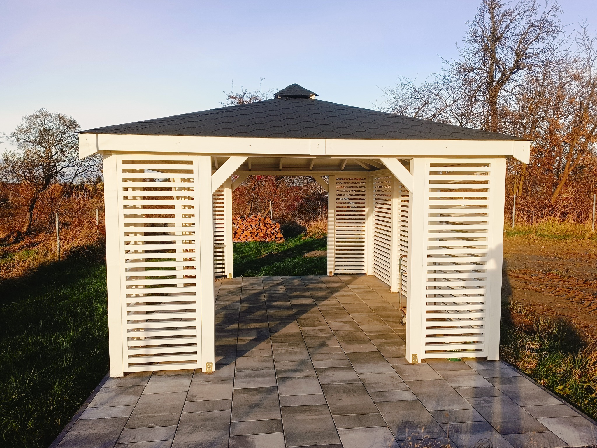 Nowoczesna, biała altana ogrodowa z czarnym dachem, otoczona zielenią i wyłożona szarą kostką brukową, z widocznym stosem drewna wewnątrz.