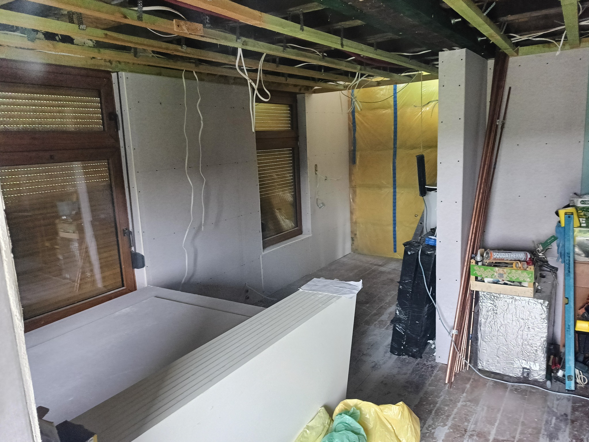 Wnętrze w trakcie prac wykończeniowych: ściany obłożone płytami gipsowo-kartonowymi, widoczne drewniane elementy konstrukcyjne sufitu, okno z żaluzjami, instalacje elektryczne i rury miedziane...