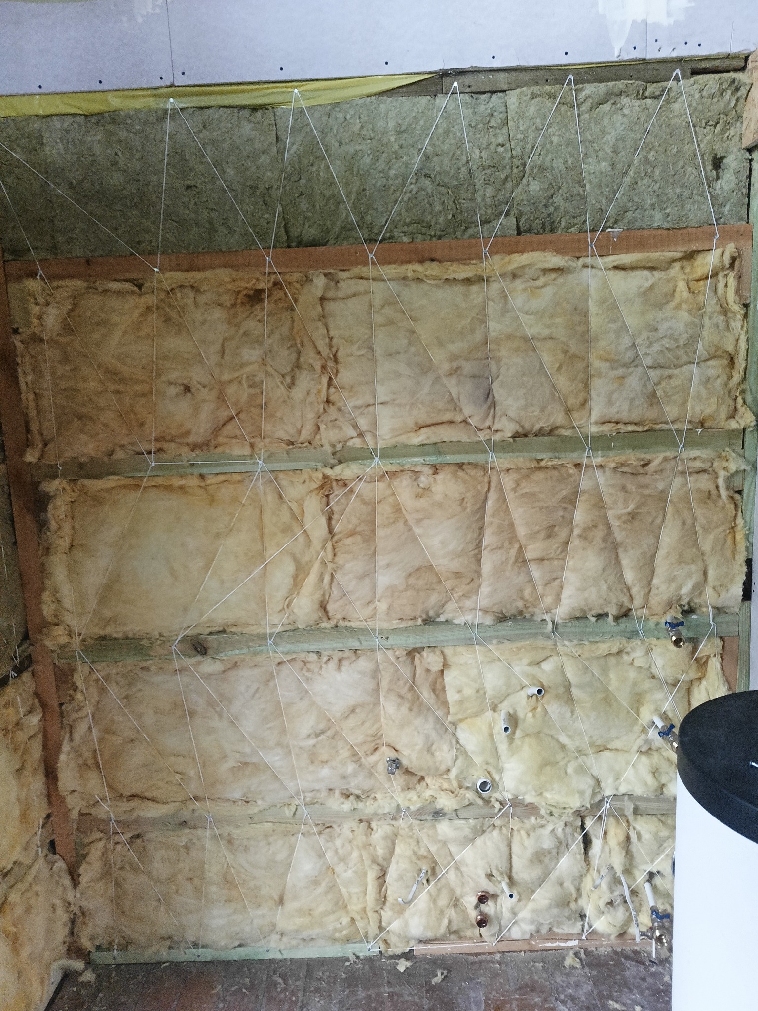 Izolacja z wełny mineralnej zamontowana na poddaszu, zabezpieczona sznurkiem, widoczne elementy instalacji wodnej.