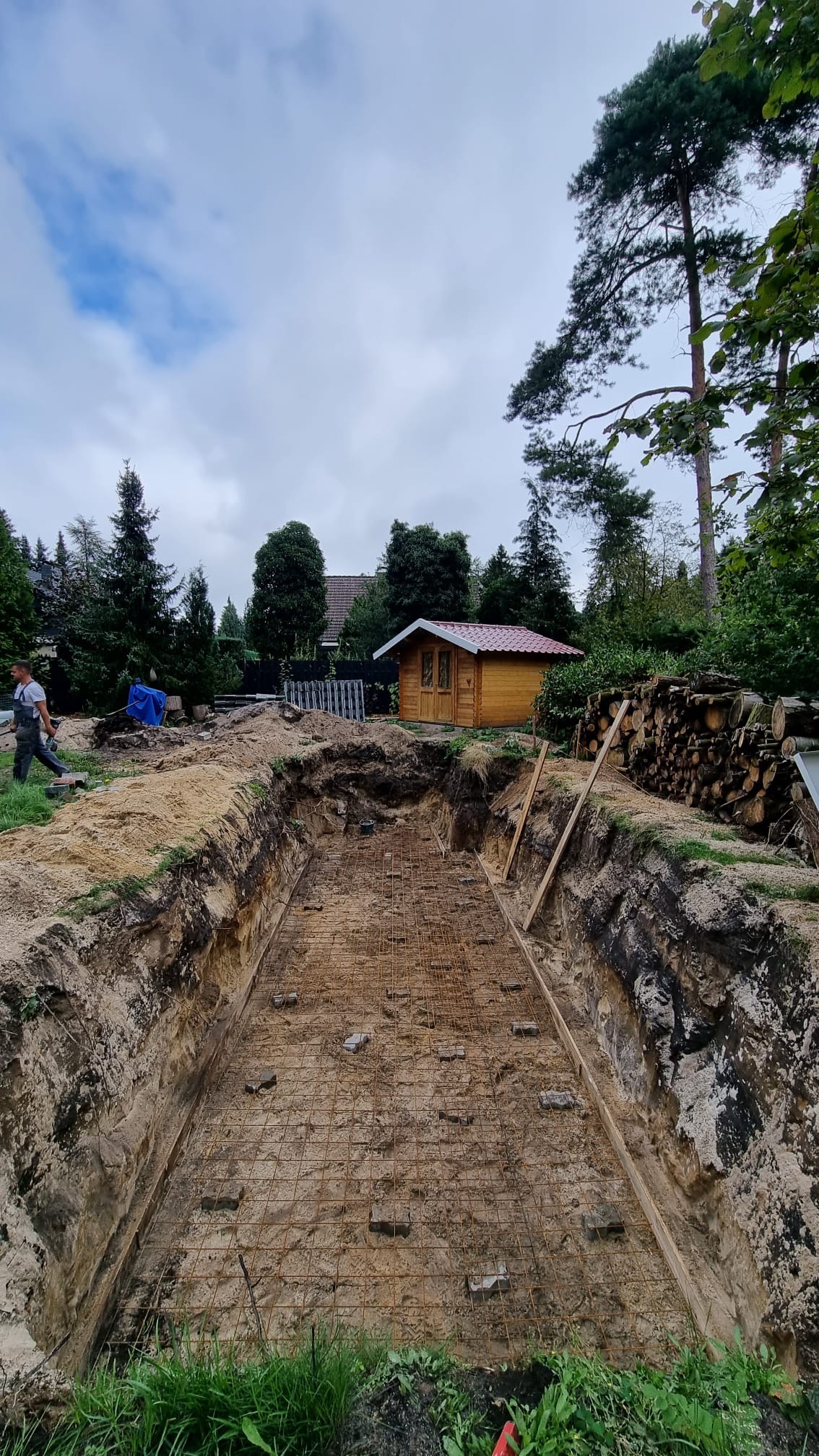 Wykop pod budowę zbiornika wodnego z ułożoną siatką zbrojeniową, widoczna drewniana altana w tle i pracownik w oddali.