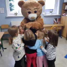 Grupa dzieci otacza pluszowego misia w klasie z biurkami i regałem z książkami, widoczny fragment flagi brytyjskiej.