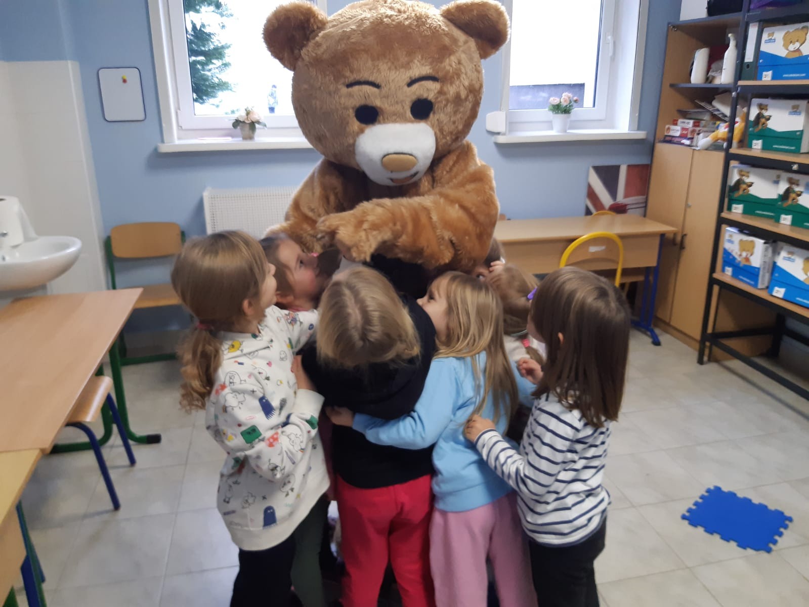 Grupa dzieci otacza pluszowego misia w klasie z biurkami i regałem z książkami, widoczny fragment flagi brytyjskiej.
