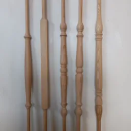 Pięć surowych, drewnianych słupków balustradowych w różnych kształtach, zawieszonych na haczykach na białej ścianie.