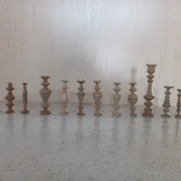 Kolekcja kilkunastu drewnianych, surowych elementów toczonych o różnych kształtach, przypominających nogi do mebli lub świeczniki, ustawionych w rzędzie na blacie.