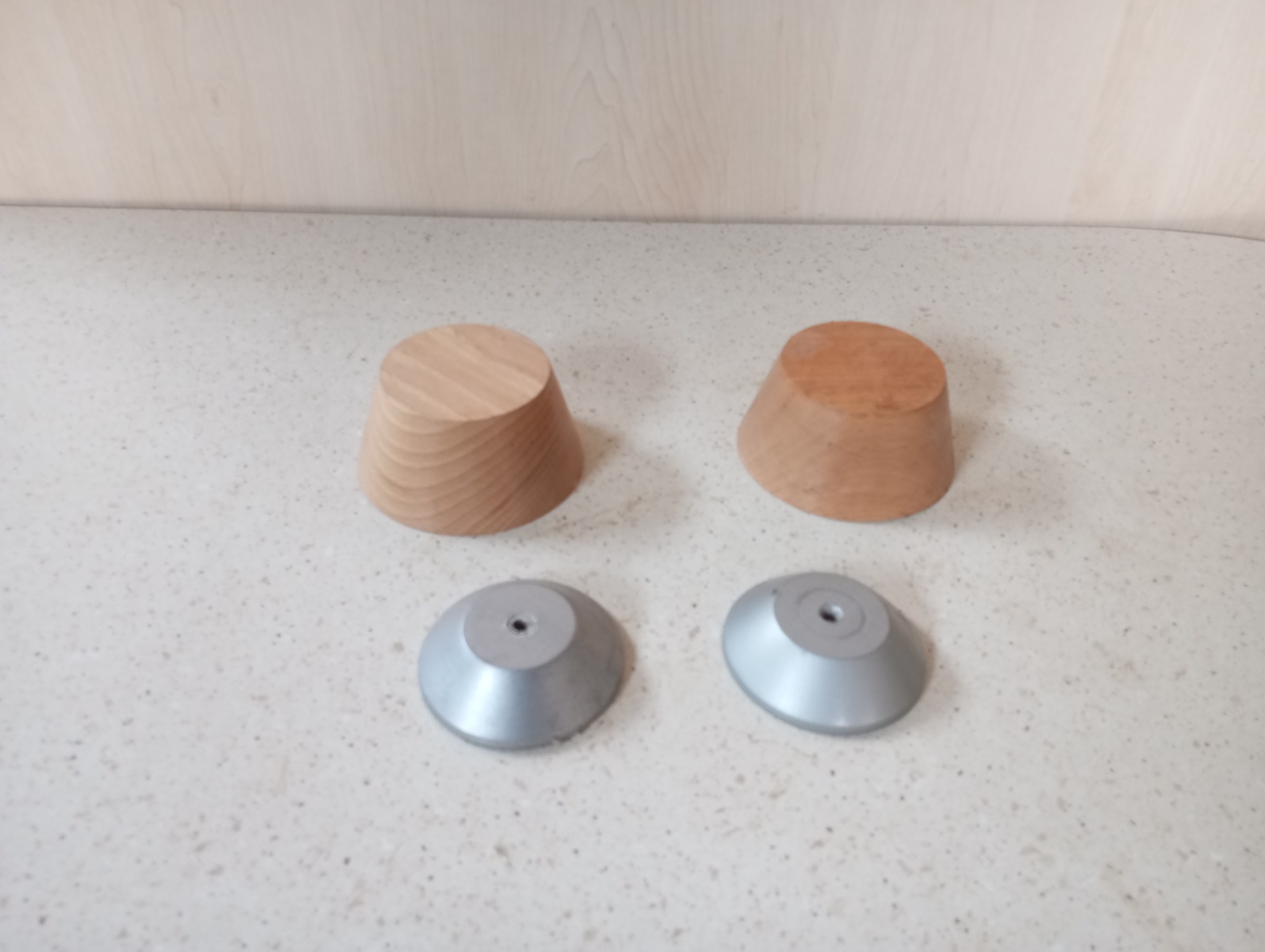 Dwa drewniane, stożkowe elementy i dwa metalowe, okrągłe podstawki z otworem na śrubę, ułożone na jasnym blacie.