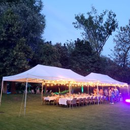 Michał Ostrowski GASTROOSTRY - Uroczyste przyjęcie na świeżym powietrzu pod białym namiotem, oświetlonym girlandami żarówek, z ustawionymi stołami i krzesłami, oświetlone dodatkowo fioletowym światłem, na tle zieleni drzew...