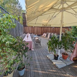 Michał Ostrowski GASTROOSTRY - Ustawiony stół z różowym obrusem na drewnianym tarasie, przygotowany do wydarzenia pod dużym parasolem z logo restauracji, otoczony roślinami w doniczkach.