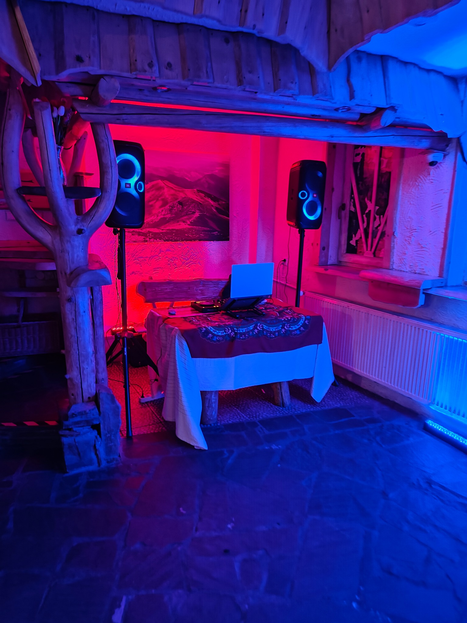 DJ setup z laptopem i głośnikami JBL oświetlony czerwonym i niebieskim światłem w rustykalnym wnętrzu.
