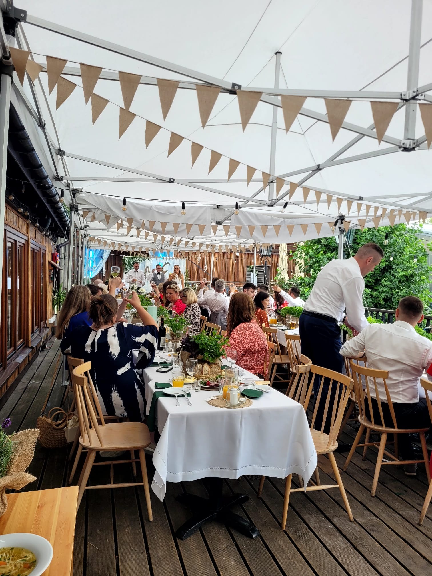 Uroczyste przyjęcie pod białym namiotem z jutowymi proporczykami, obsługa kelnerska, drewniany taras, goście przy stołach z białymi obrusami i zielonymi akcentami, dekoracje roślinne.