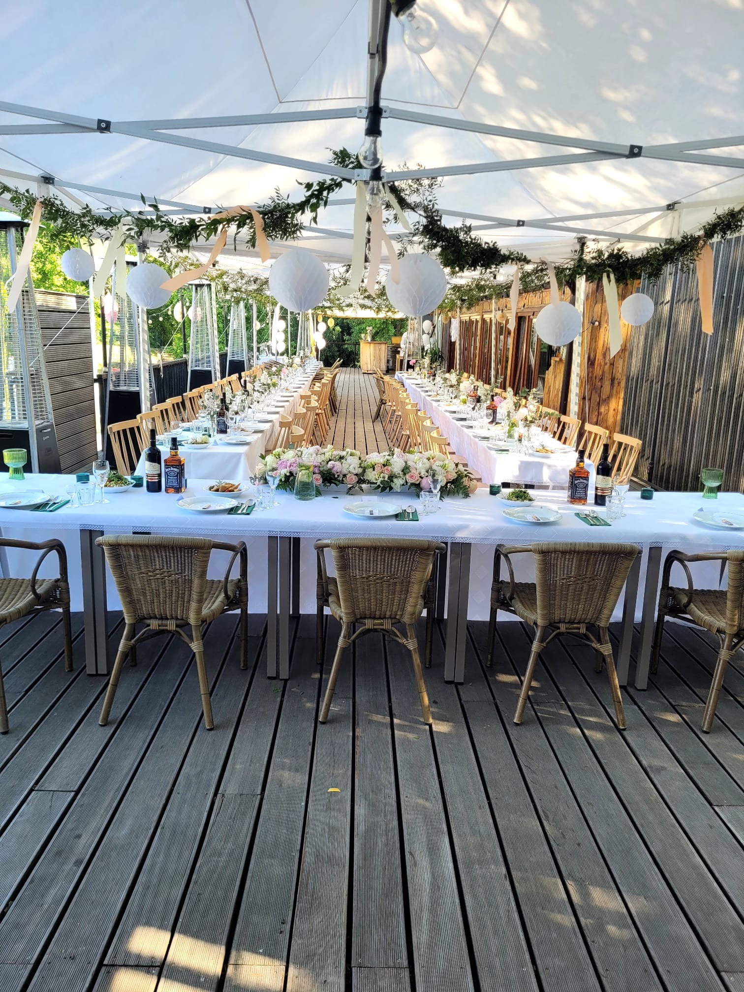 Długi stół nakryty białym obrusem, udekorowany kompozycjami kwiatowymi i butelkami alkoholu, ustawiony na drewnianym tarasie pod białym namiotem z dekoracjami z zieleni i papierowych lampionów.
