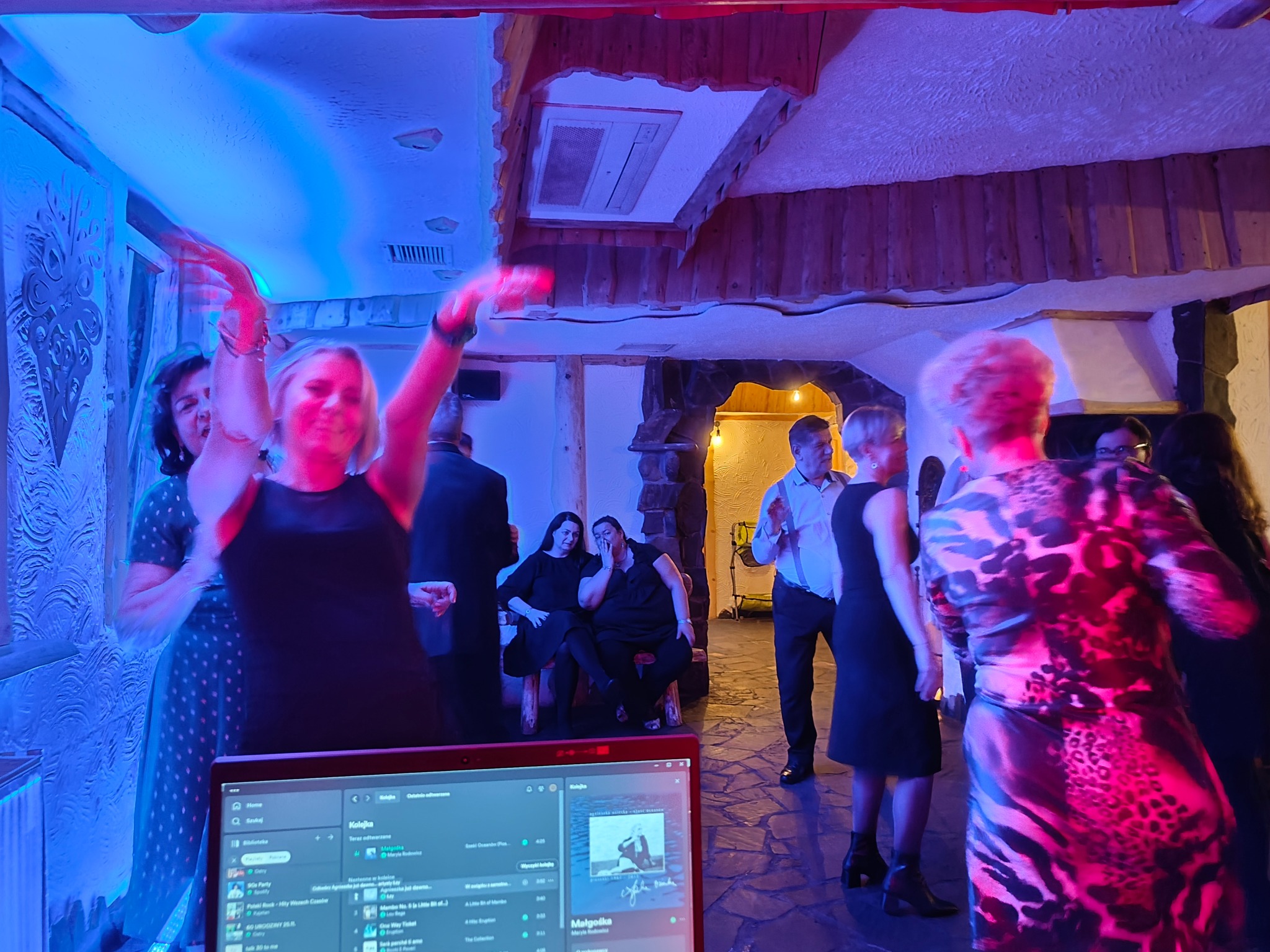 Roztańczone osoby na imprezie, widoczny laptop z listą odtwarzania muzyki, dekoracyjne oświetlenie w kolorze niebieskim i fioletowym, drewniane elementy wystroju.
