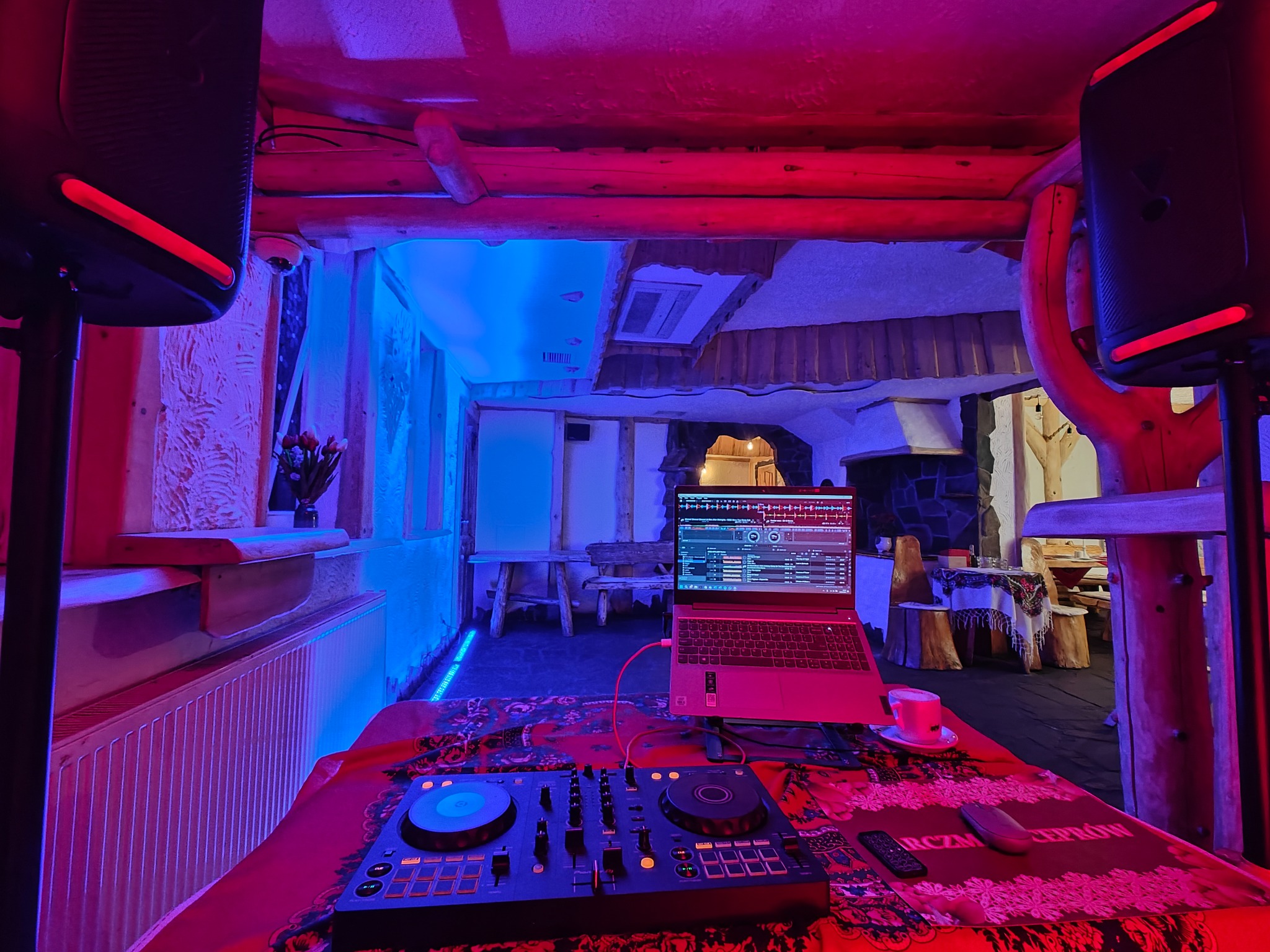 DJ set z laptopem i mikserem w rustykalnym wnętrzu z drewnianymi elementami, oświetlonym czerwono-niebieskim światłem, widoczne głośniki po bokach i stolik z filiżanką.