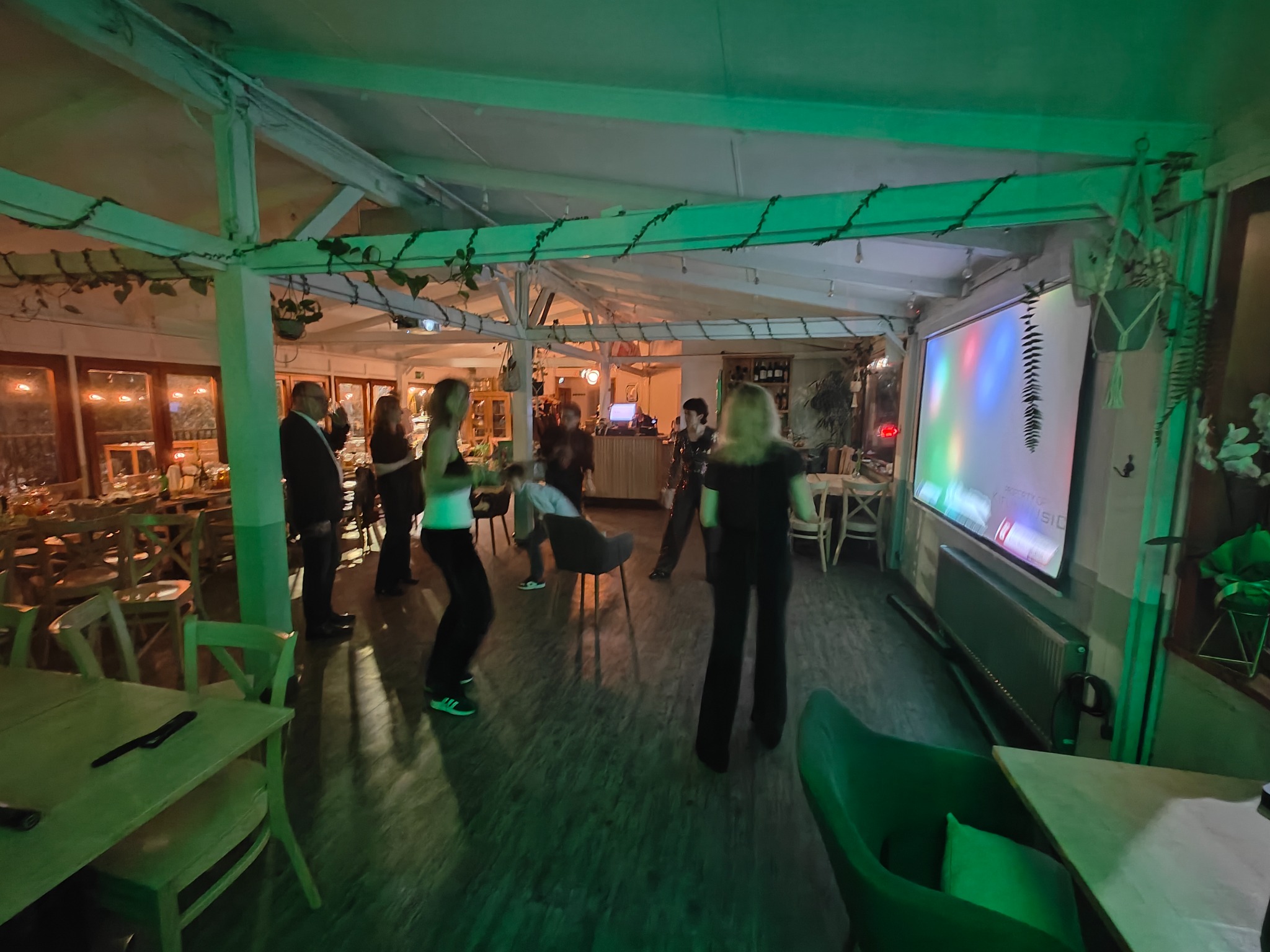 Wnętrze sali z drewnianą konstrukcją sufitu, gdzie grupa osób tańczy i ogląda projekcję na ekranie, oświetlona zielonym światłem.