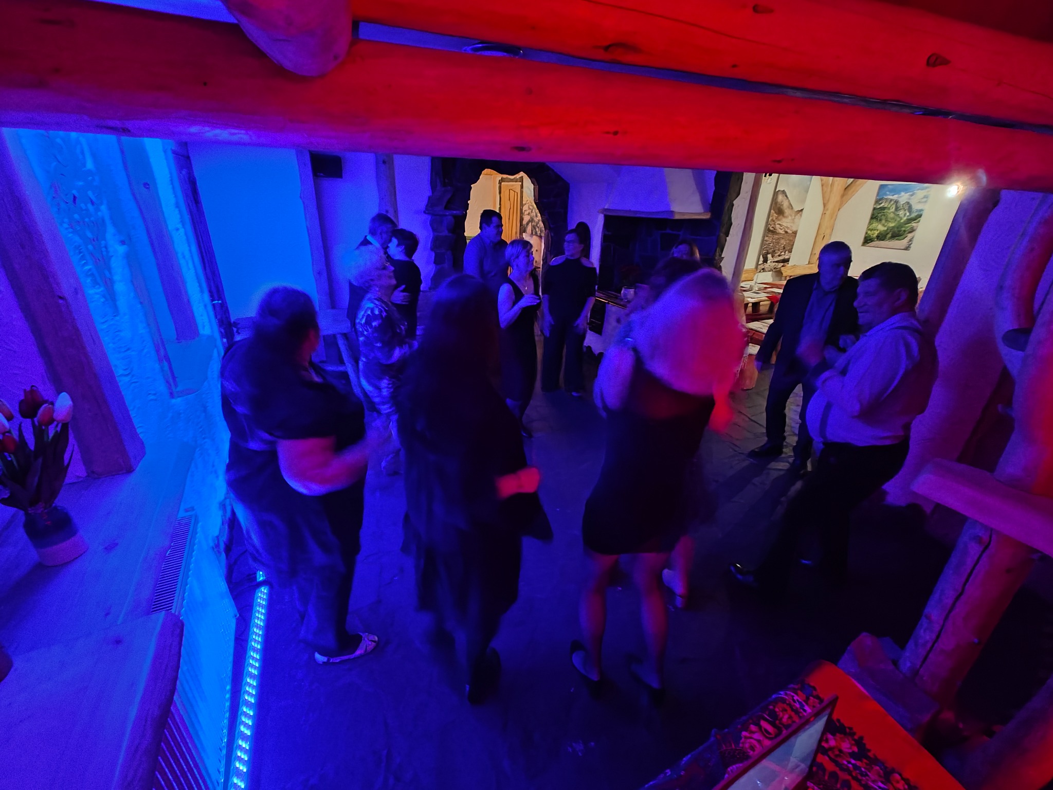 Grupa osób w pomieszczeniu z drewnianymi elementami, oświetlonym na niebiesko i czerwono, sugerującym imprezę lub wydarzenie.