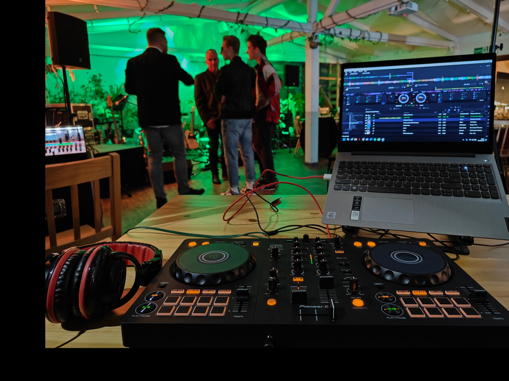Stanowisko DJ-a z mikserem Pioneer DJ, słuchawkami z czerwonymi akcentami i laptopem z interfejsem oprogramowania DJ-skiego, w tle grupa osób i sprzęt nagłośnieniowy pod zielonym oświetleniem.