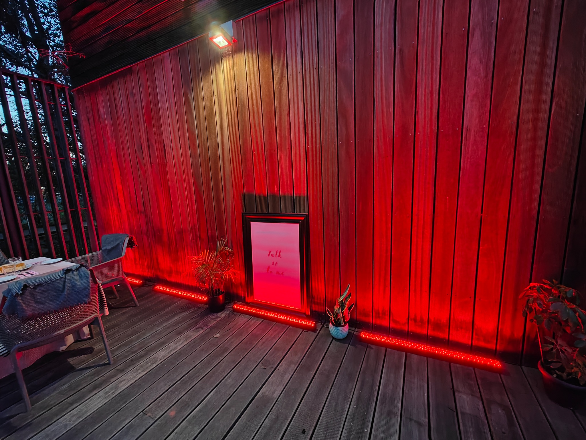 Ustawiony stół z krzesłami na drewnianym tarasie, oświetlony czerwonymi lampami LED, z drewnianą ścianą w tle i roślinami doniczkowymi.