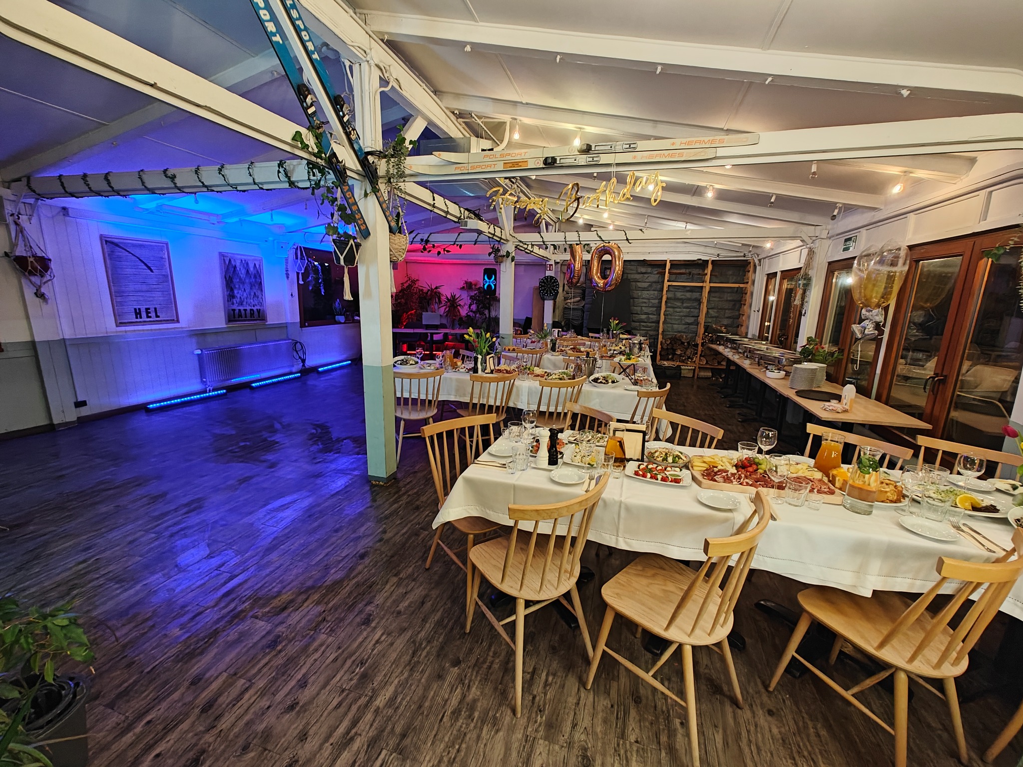 Sala bankietowa z nakrytymi stołami, drewnianymi krzesłami, dekoracjami urodzinowymi i niebieskim oświetleniem akcentującym.