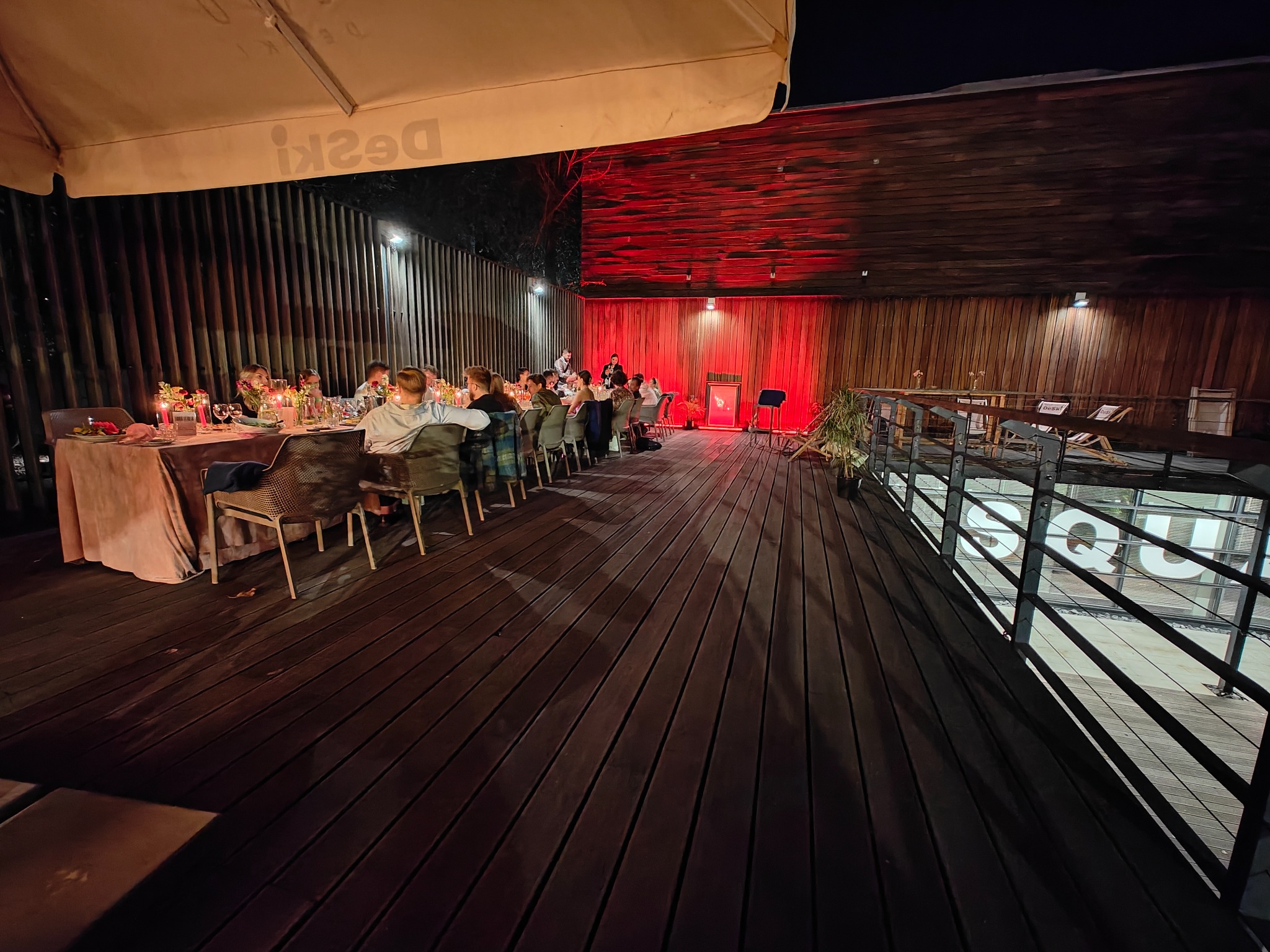 Uroczysta kolacja firmowa na drewnianym tarasie, oświetlona czerwonymi światłami, z muzyką na żywo w tle.