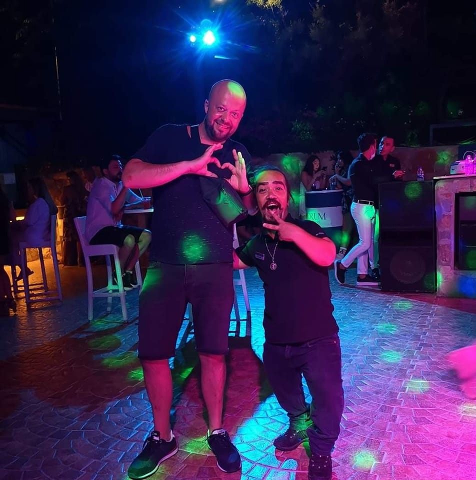 Dwóch mężczyzn pozuje na tle baru z DJ-em, oświetlonych kolorowymi światłami dyskotekowymi.