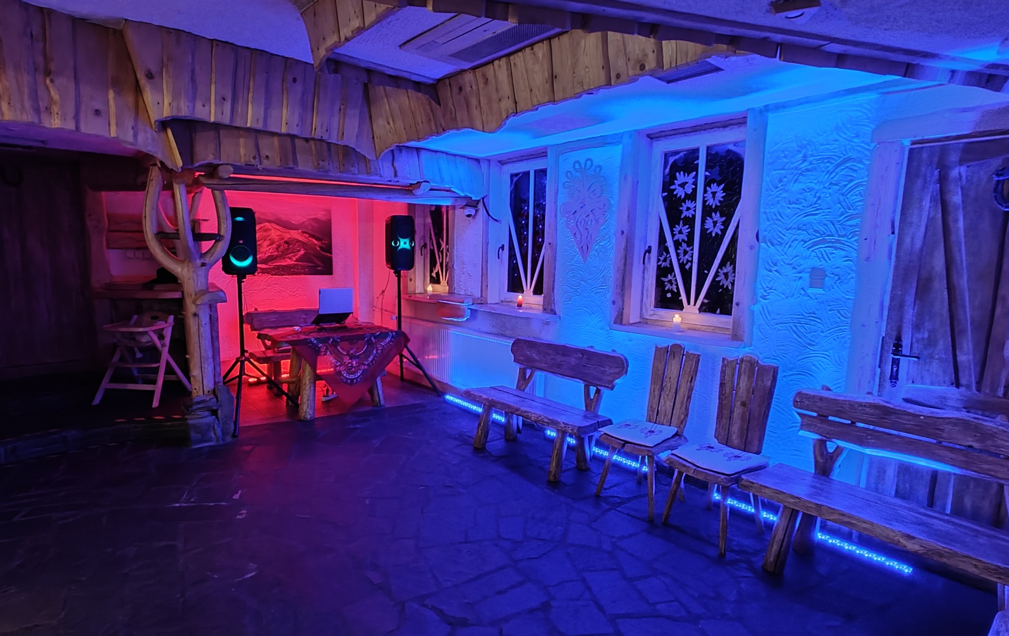 Rustykalne wnętrze z drewnianymi elementami oświetlone czerwonym i niebieskim światłem, z nagłośnieniem i laptopem na stole.