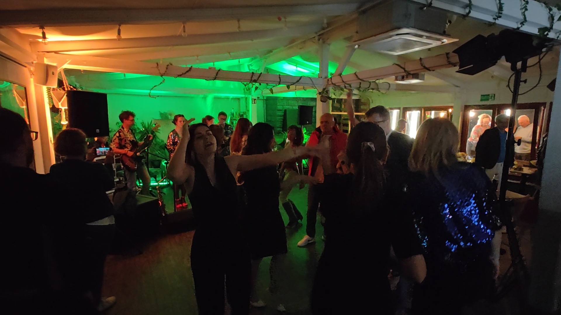 Zespół muzyczny grający na imprezie, ludzie tańczą, oświetlenie sceniczne w kolorze zielonym.