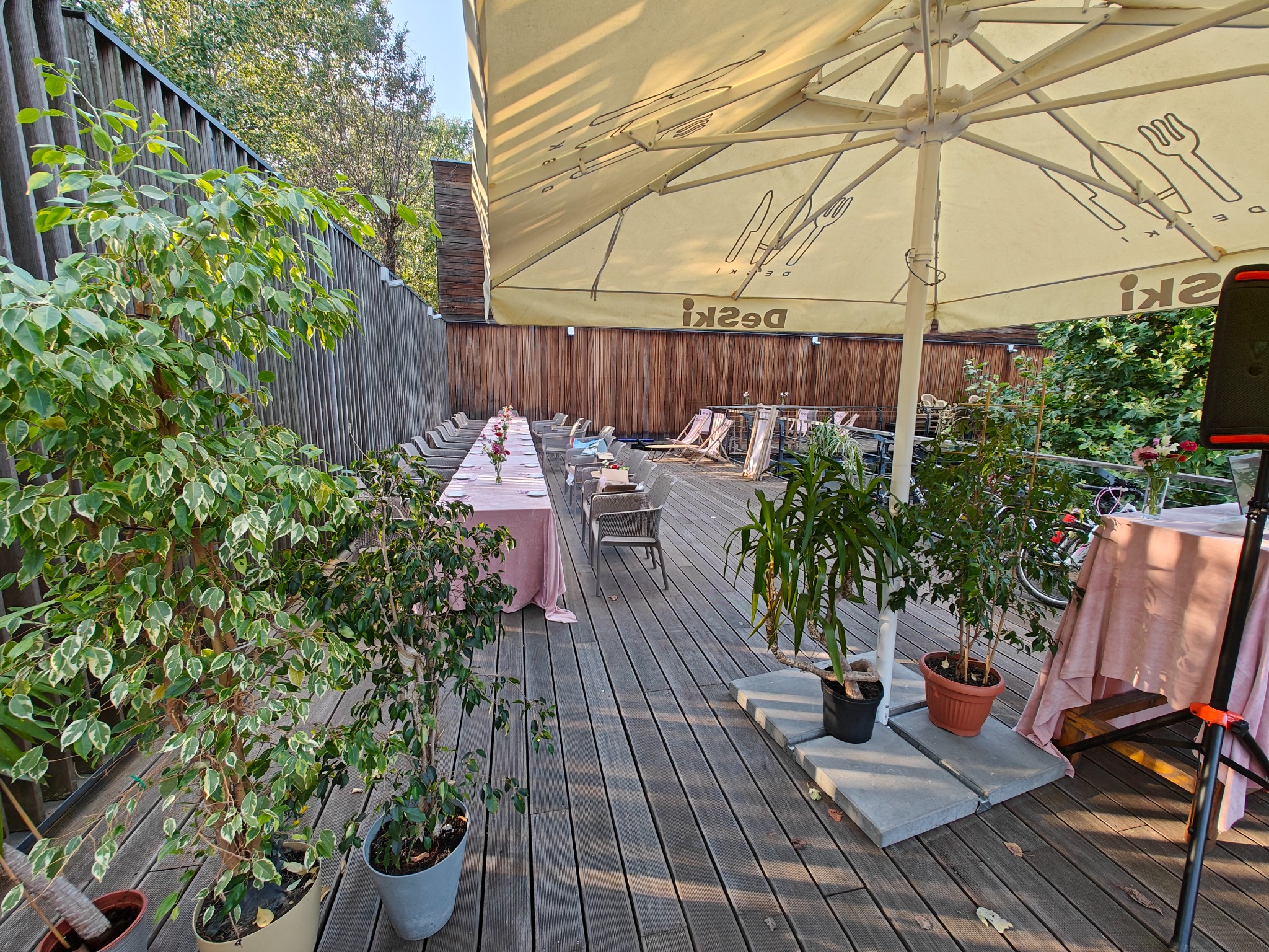 Ustawiony stół z różowym obrusem na drewnianym tarasie, przygotowany do wydarzenia pod dużym parasolem z logo restauracji, otoczony roślinami w doniczkach.