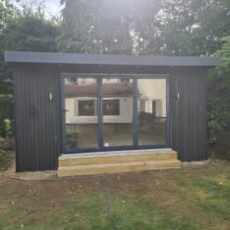 Konrad Ochnio - Nowoczesna, grafitowa altana ogrodowa z przesuwnymi szklanymi drzwiami i drewnianymi schodami wejściowymi, otoczona zielenią.