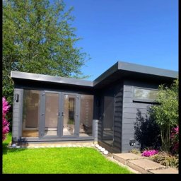 Konrad Ochnio - Nowoczesny, szary domek ogrodowy z dużymi oknami i drzwiami, otoczony zielonym trawnikiem i kwitnącymi krzewami w słoneczny dzień.