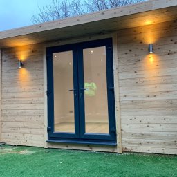 Konrad Ochnio - Nowoczesny domek ogrodowy z drewnianą elewacją, oświetleniem zewnętrznym i dwuskrzydłowymi drzwiami w kolorze antracytowym, widok z zewnątrz na sztuczną trawę.