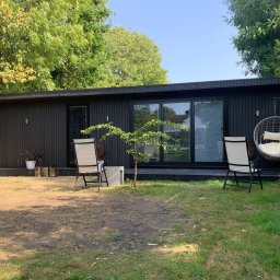 Konrad Ochnio - Nowoczesny, ciemny domek letniskowy z dużymi oknami przesuwnymi, otoczony zielenią, z wiszącym fotelem i krzesłami na zewnątrz, z widocznym fragmentem trawnika.