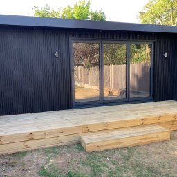 Konrad Ochnio - Nowoczesna, czarna altana z przesuwnymi szklanymi drzwiami i drewnianym tarasem z dwoma stopniami, na trawniku.