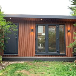 Konrad Ochnio - Nowoczesny domek ogrodowy z brązową elewacją z pionowych paneli, szare okna i drzwi, oświetlenie zewnętrzne, otoczony zielenią.