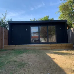 Nowoczesny, czarny domek ogrodowy z przesuwnymi drzwiami i drewnianym podestem, otoczony trawnikiem i drewnianym ogrodzeniem.