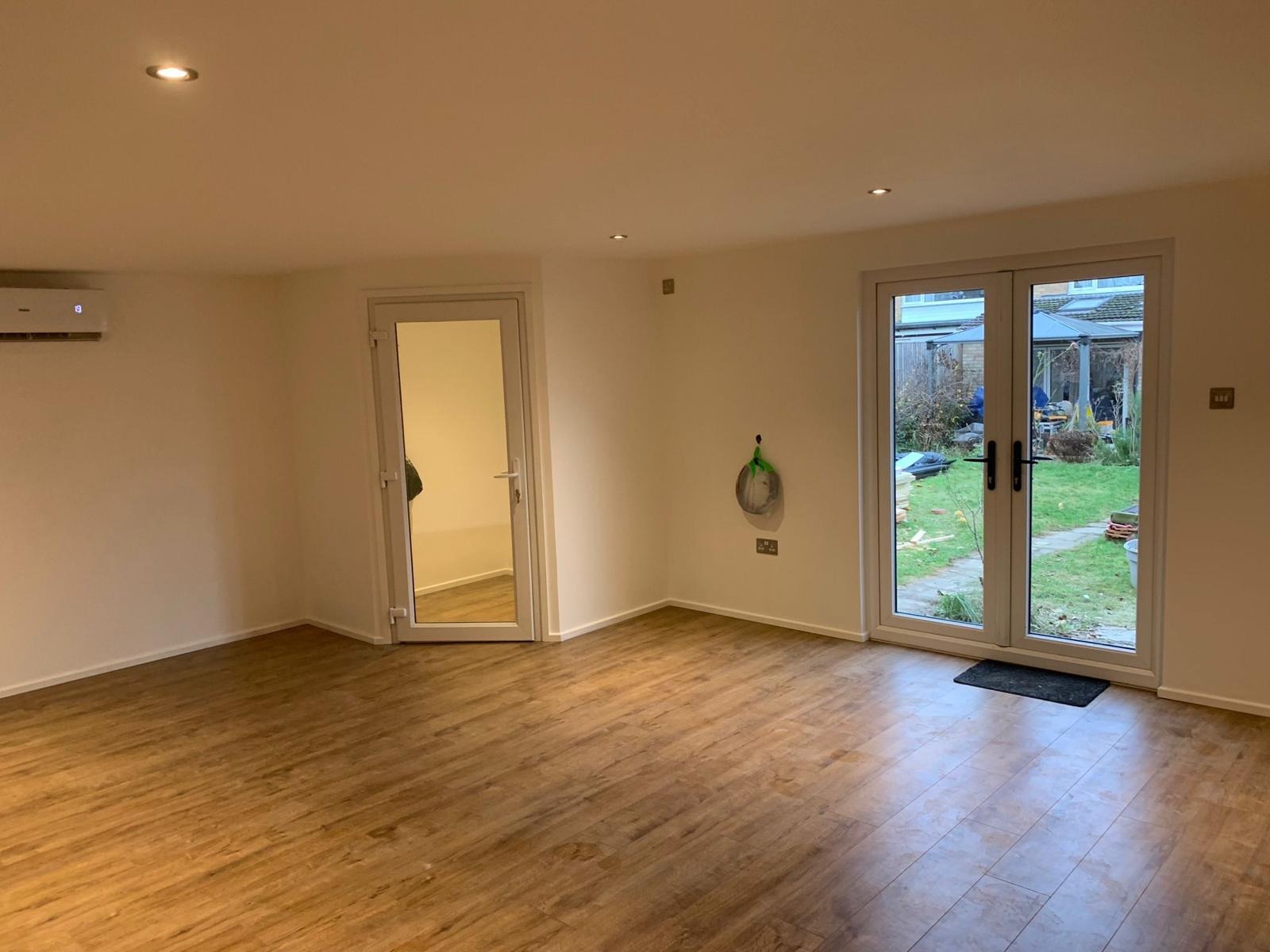 Wykończone wnętrze z panelami podłogowymi, białymi ścianami, klimatyzacją i dwuskrzydłowymi drzwiami balkonowymi wychodzącymi na ogród.