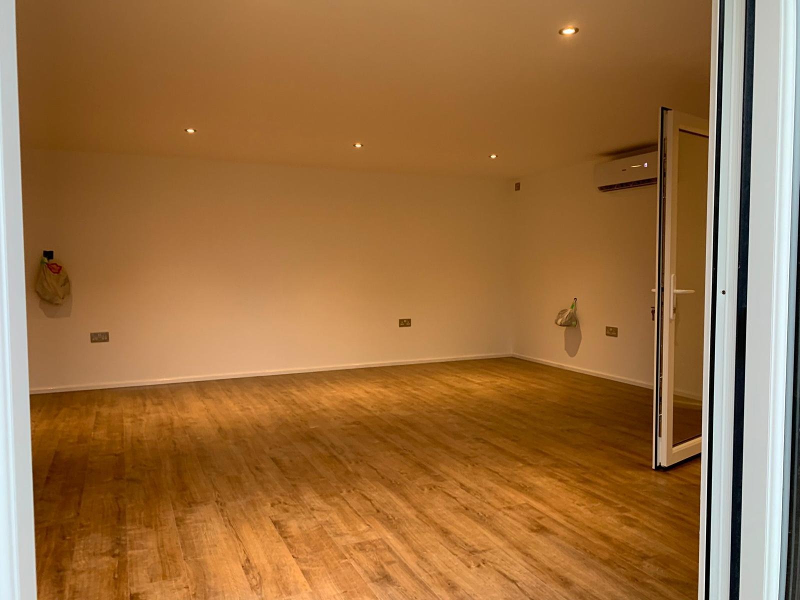 Puste, jasne pomieszczenie z panelami podłogowymi imitującymi drewno, widok zza otwartych drzwi balkonowych, klimatyzacja na ścianie, oświetlenie punktowe w suficie.