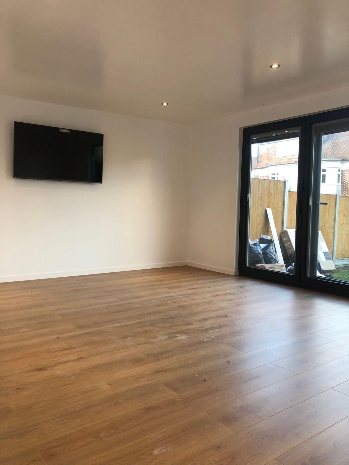 Jasne pomieszczenie z panelami podłogowymi w odcieniach brązu, białymi ścianami, zamontowanym telewizorem i oknem balkonowym z widokiem na ogród, częściowo zasłonięte workami i elementami po remoncie.