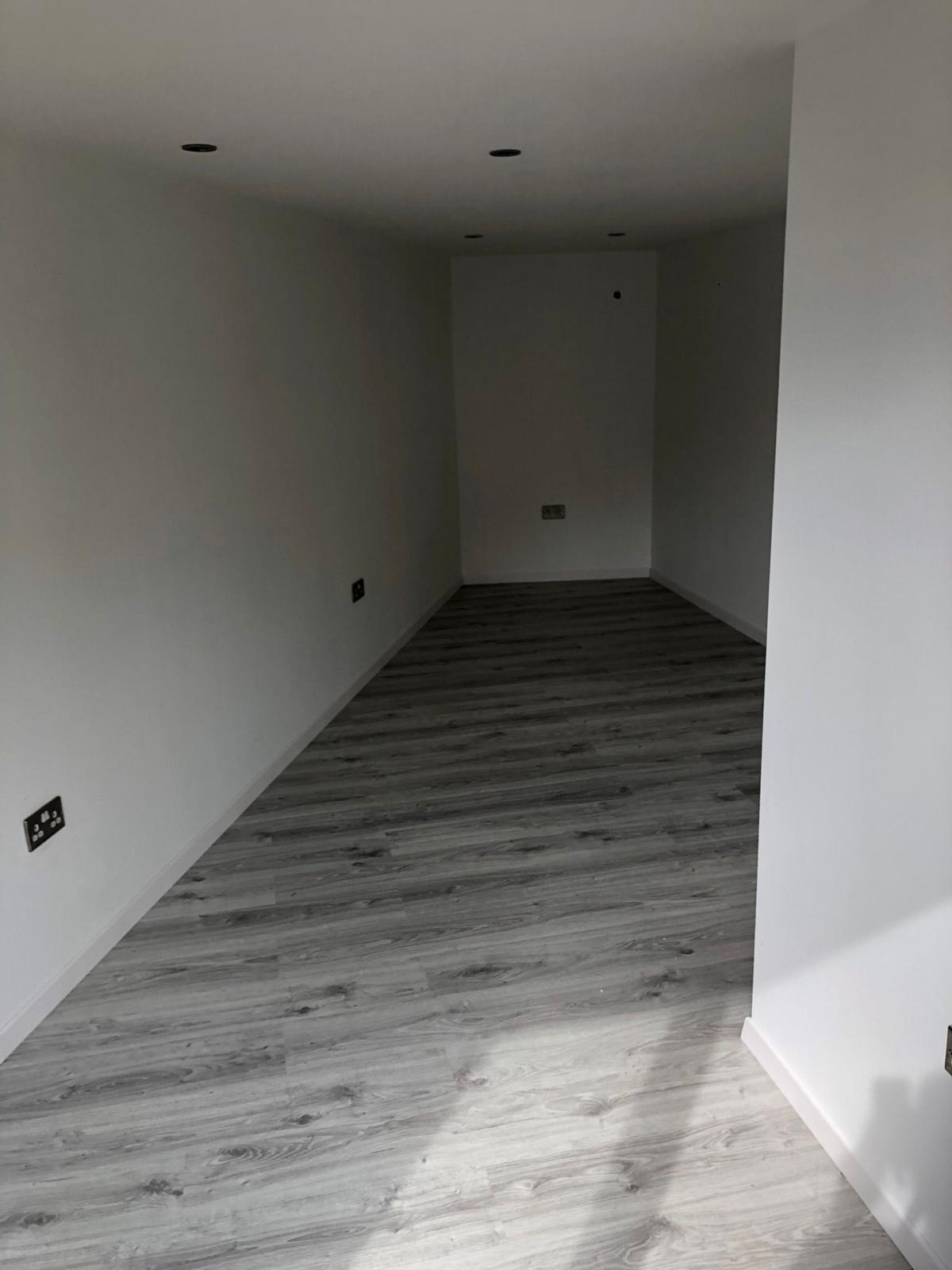Wnętrze pomieszczenia po remoncie: szare panele podłogowe imitujące drewno, świeżo pomalowane białe ściany z gniazdkami elektrycznymi, czarne punkty oświetleniowe w suficie.
