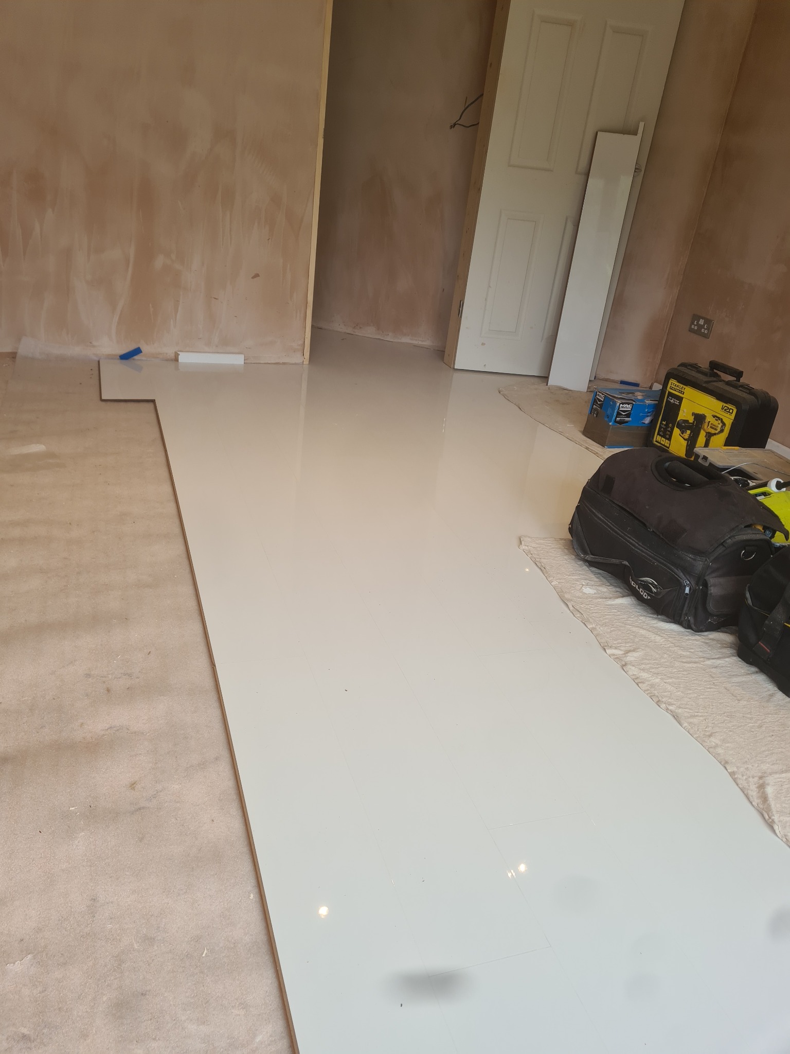 Fragment podłogi w trakcie prac: ułożone białe panele podłogowe kontrastują z surowym, nieprzygotowanym podłożem; widoczne narzędzia i torby monterskie w tle.