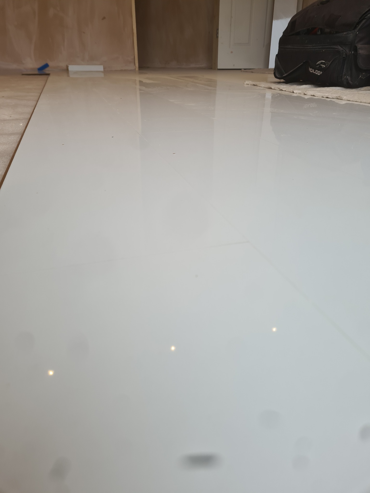 Błyszcząca, biała podłoga z płytek ceramicznych po remoncie, widoczna torba narzędziowa w tle oraz narzędzia do układania płytek przy ścianie.