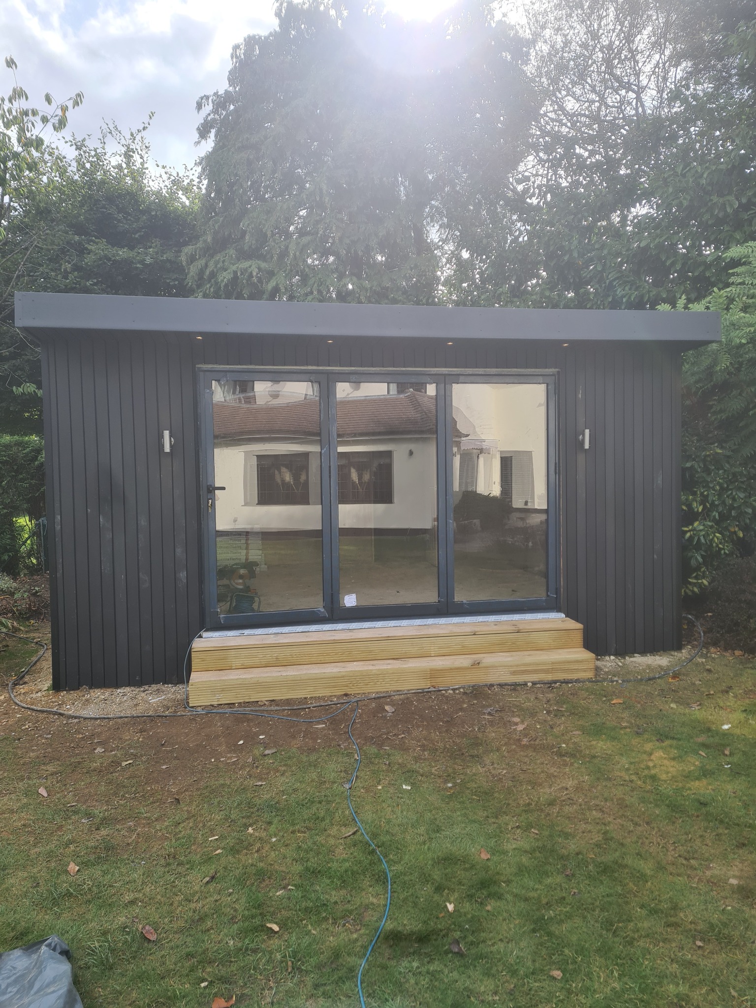 Nowoczesna, grafitowa altana ogrodowa z przesuwnymi szklanymi drzwiami i drewnianymi schodami wejściowymi, otoczona zielenią.